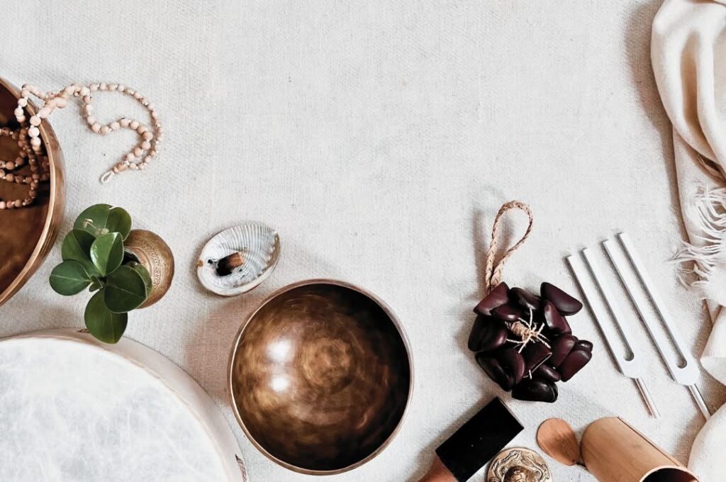 A collection of sound healing instruments arranged on a white natural wool cloth: seed rattles, a tulsi meditation mala, two tuning forks, a shamanic drum, and palo santo.